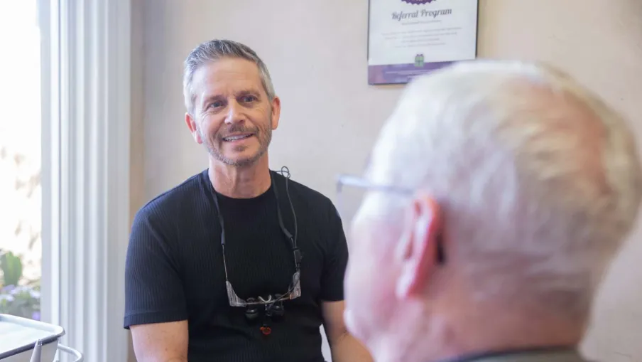 Dentist smiling and talking with elderly male patient in a bright dental office near window.