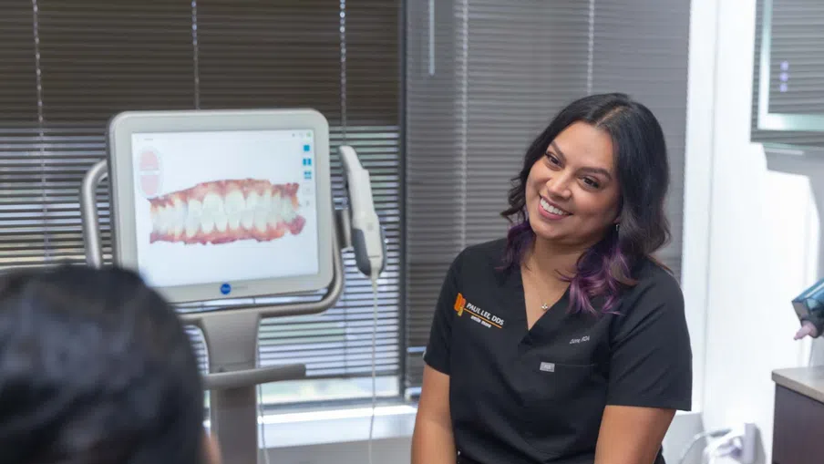 Smiling dental professional in black scrubs consulting patient with dental x-ray on screen in clinic.