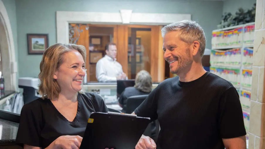 Smiling healthcare worker showing a digital tablet to a male patient in a medical office reception area.
