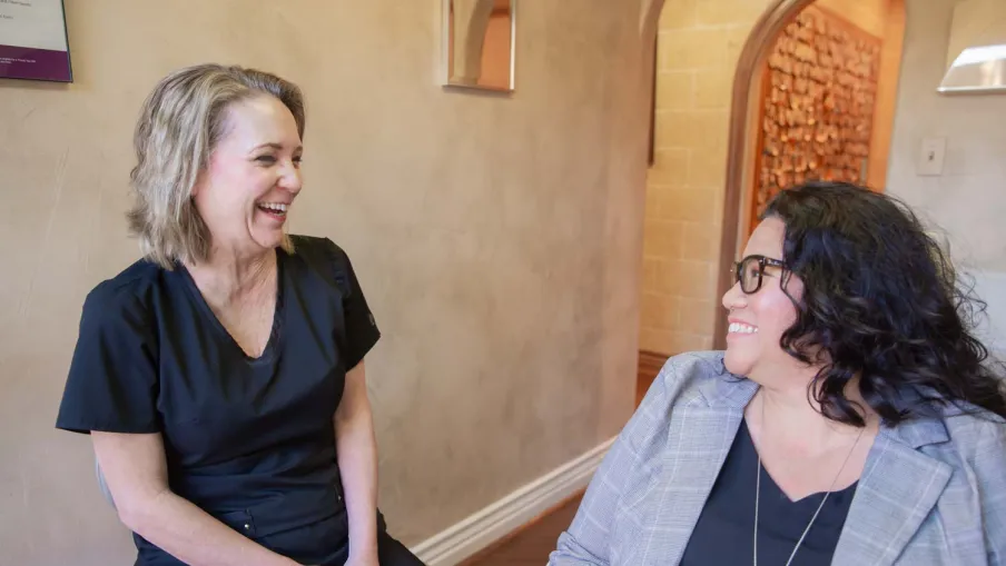 Two women smiling and chatting in a warm, casual indoor setting with neutral walls and wooden floors.
