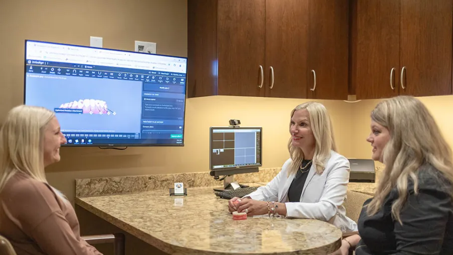 Dentist and assistant consulting patient with dental model and digital teeth imaging on screens in exam room.