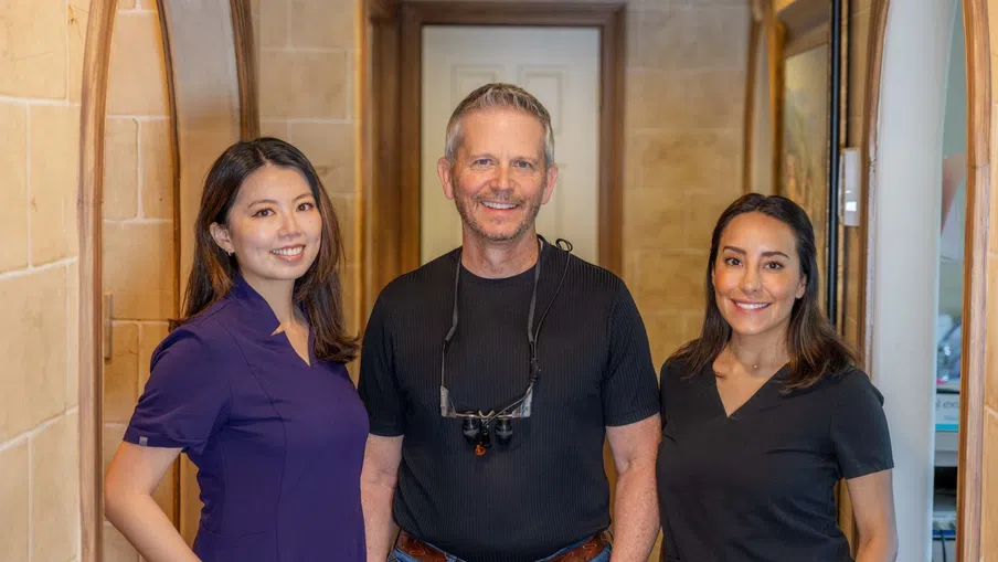 Three dental professionals in scrubs and casual attire smile in a warmly lit hallway with tiled walls and arched doorways.