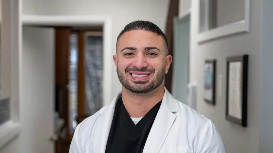 Smiling male doctor in white coat with black scrubs standing in a medical office hallway.