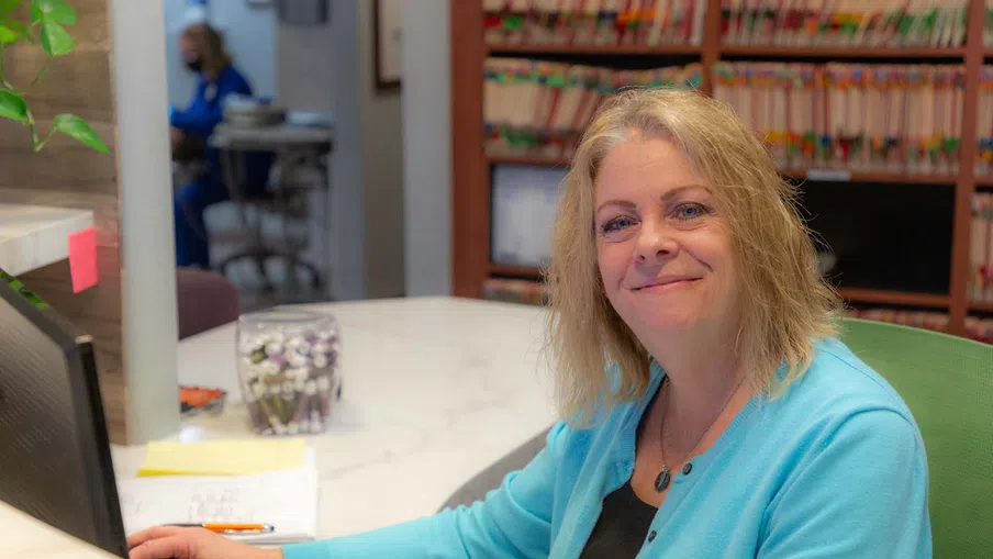 Smiling woman in a blue cardigan working at a reception desk with medical files in the background.