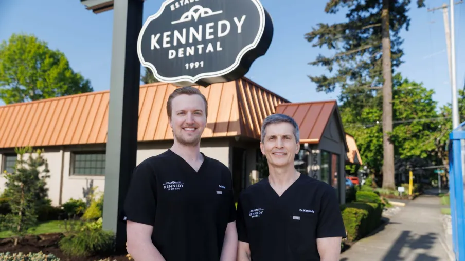 Two dentists in black Kennedy Dental scrubs standing outside their dental clinic under the clinic sign on a sunny day