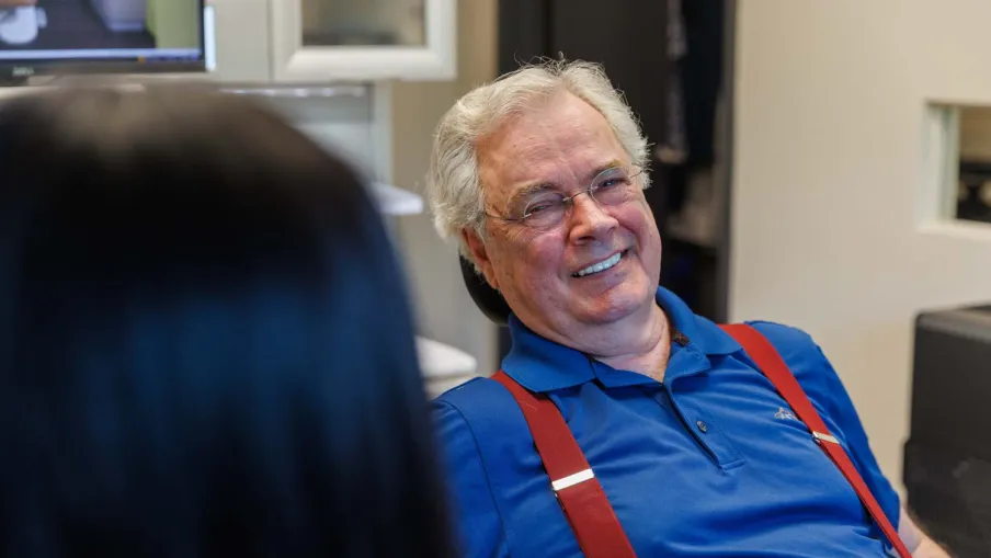 Senior man with white hair and glasses smiling, wearing blue polo and red suspenders indoors with blurred person in foreground.
