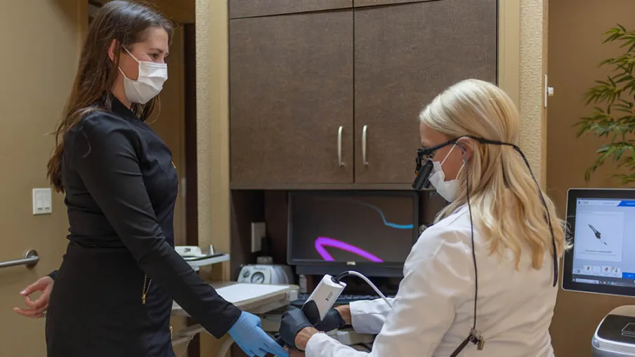 Dental professional using a handheld scanner on patient's teeth while assistant observes in clinic room.