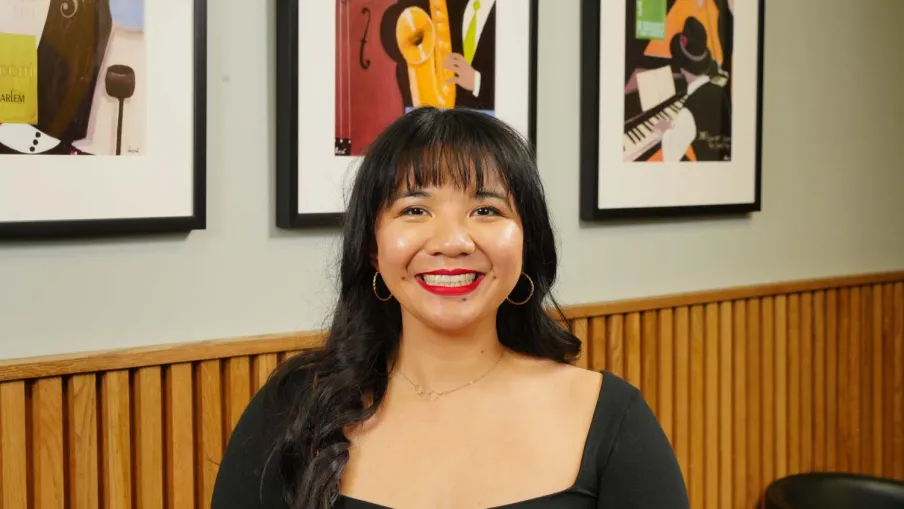 Smiling woman with long dark hair and hoop earrings sits indoors with framed art and wooden paneling behind her