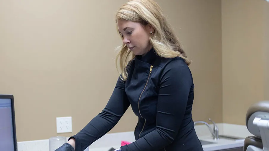 Dental professional wearing black gloves preparing tools in a clinical setting with beige walls and a computer screen.