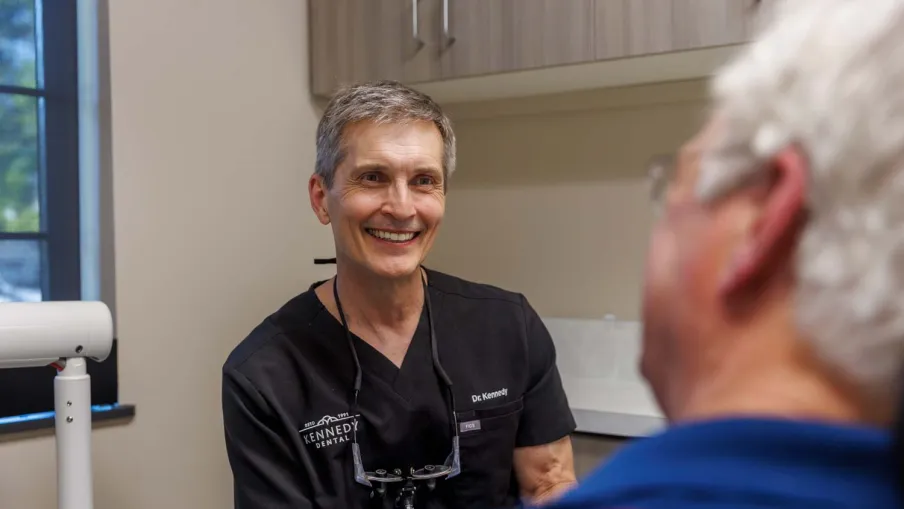 Smiling male dentist in black scrubs consulting with an elderly patient in a dental clinic room.