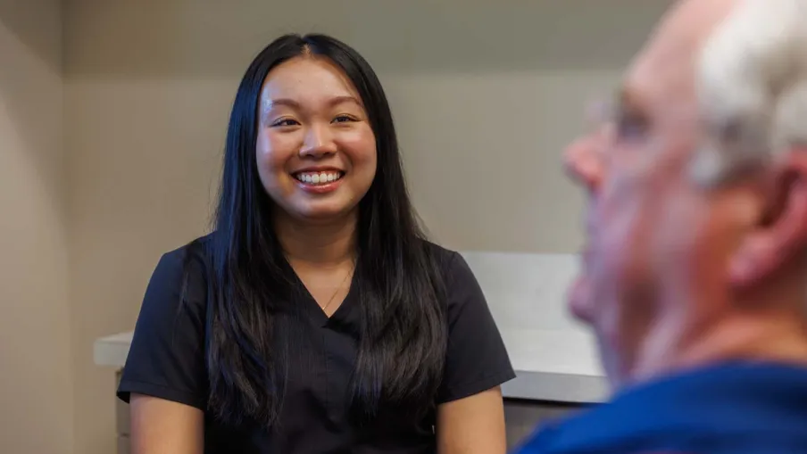 Smiling female healthcare worker in black scrubs talking to elderly male patient in clinic room.