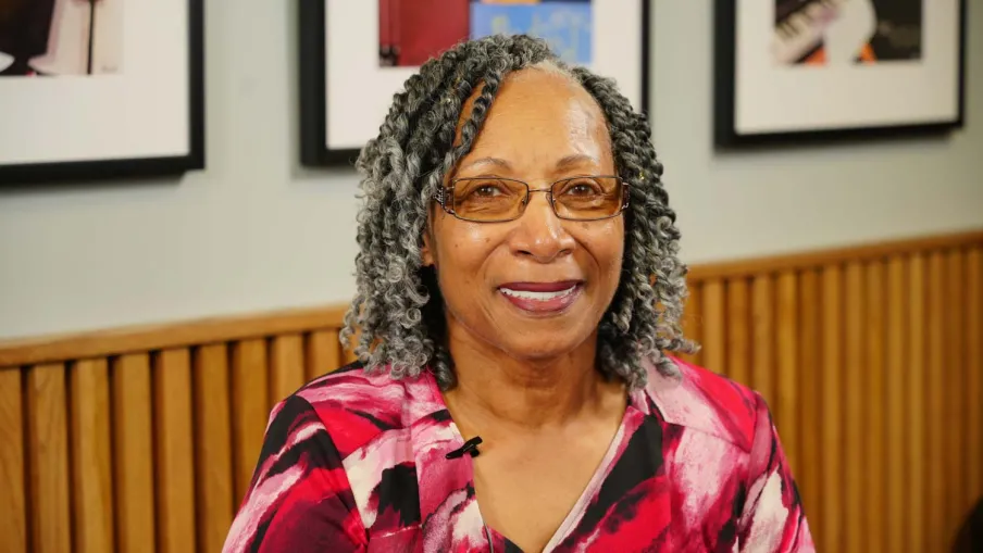 Smiling older woman with gray curly hair and glasses wearing a red patterned blouse indoors.