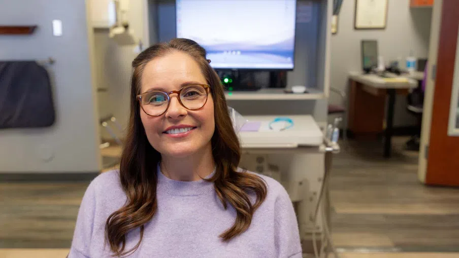 Smiling woman with glasses and purple sweater sitting in an office with computer and medical equipment.