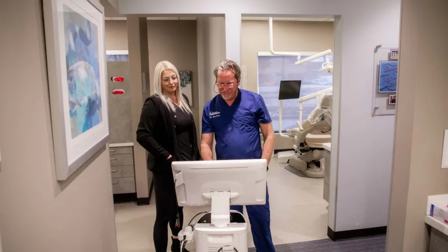 Dentist in blue scrubs and woman review digital images on a monitor in a modern dental clinic hallway.