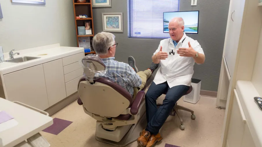 Dentist in white coat consulting with elderly male patient seated in dental chair inside clinic room.