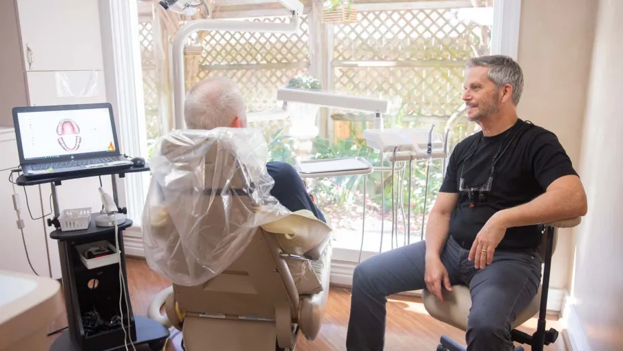 Dentist consulting an elderly patient in a bright dental office with digital dental chart on laptop screen.