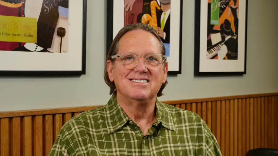 Middle-aged man wearing glasses and a green plaid shirt sitting indoors with framed musical art behind him