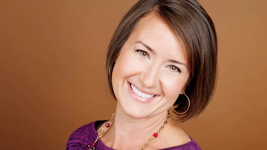 Smiling woman with short brown hair wearing a purple top and gold hoop earrings against a brown background