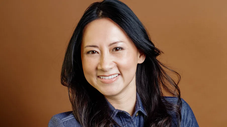 Smiling woman with long black hair wearing a blue denim shirt against a brown background.