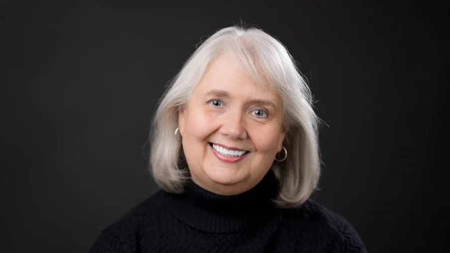 Smiling mature woman with white hair wearing black turtleneck and hoop earrings against dark background