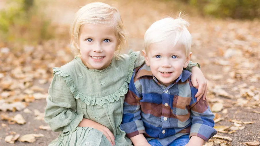 Two young children, a girl in a green dress and a boy in a plaid shirt, sitting on a leaf-strewn path smiling.