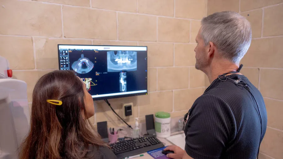 Dentist and patient reviewing dental X-rays on computer screen in clinic with beige tiled walls.