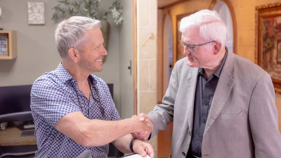 Two elderly men smiling and shaking hands indoors in a warmly lit room with decor in background