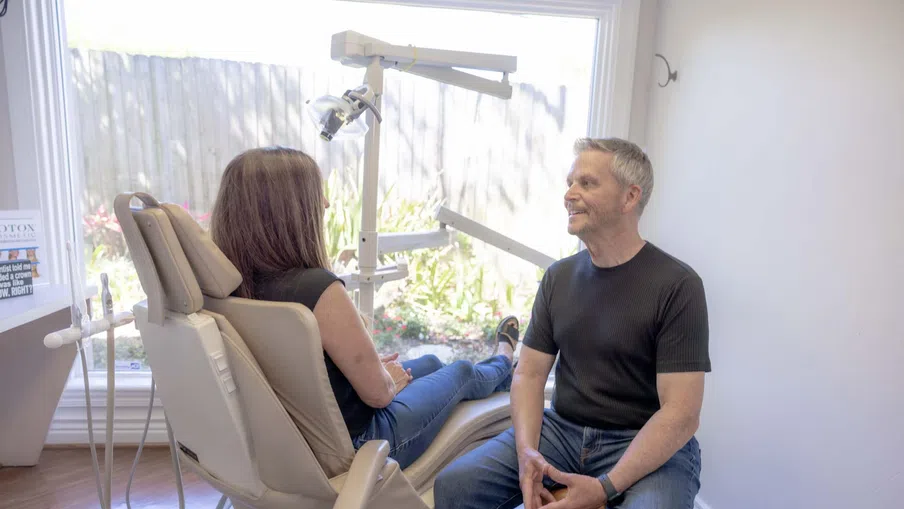 Dentist and patient interacting in a bright dental office with equipment and a large window showing plants outside.