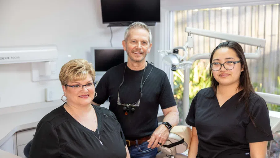 Three dental professionals in a modern clinic smiling at the camera, wearing black scrubs and dental equipment.