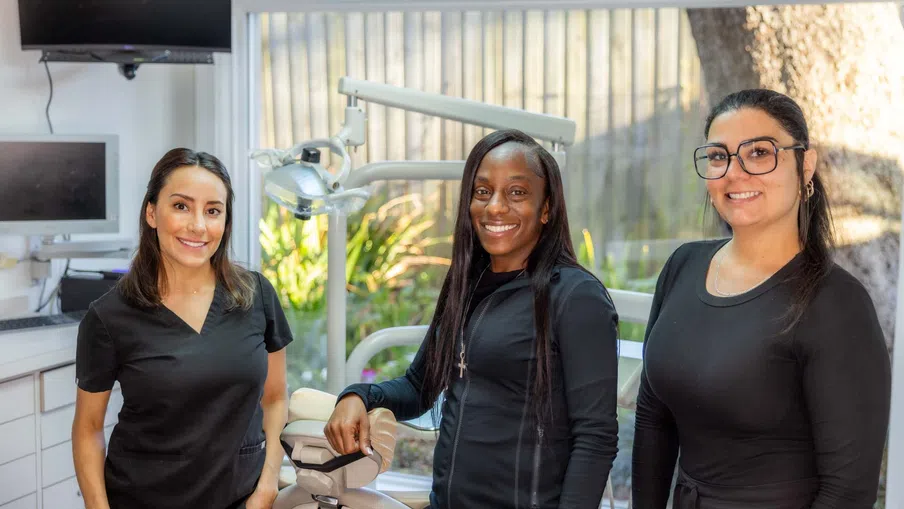 Three smiling dental professionals standing in a modern dental clinic with equipment and monitors.