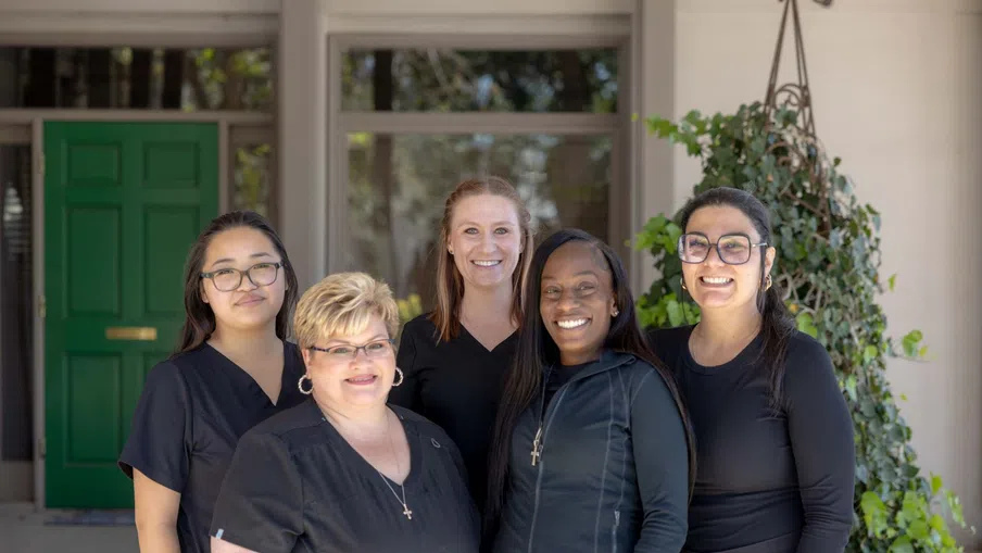 Five diverse smiling women standing outside a building with a green door, dressed in black clothing.