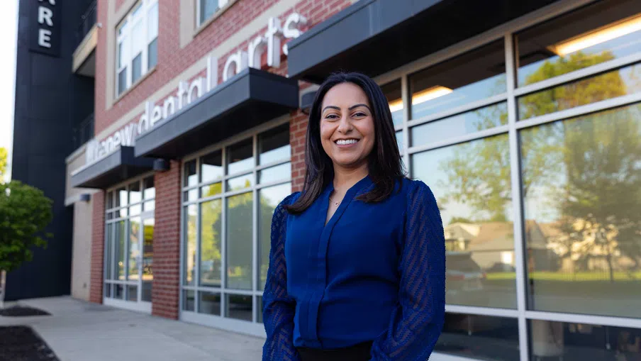 Smiling woman in a blue blouse standing confidently outside a modern dental clinic building in daytime.