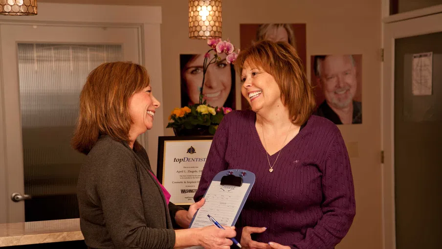 Two women smiling and discussing over a clipboard in a warmly lit dental office reception area.