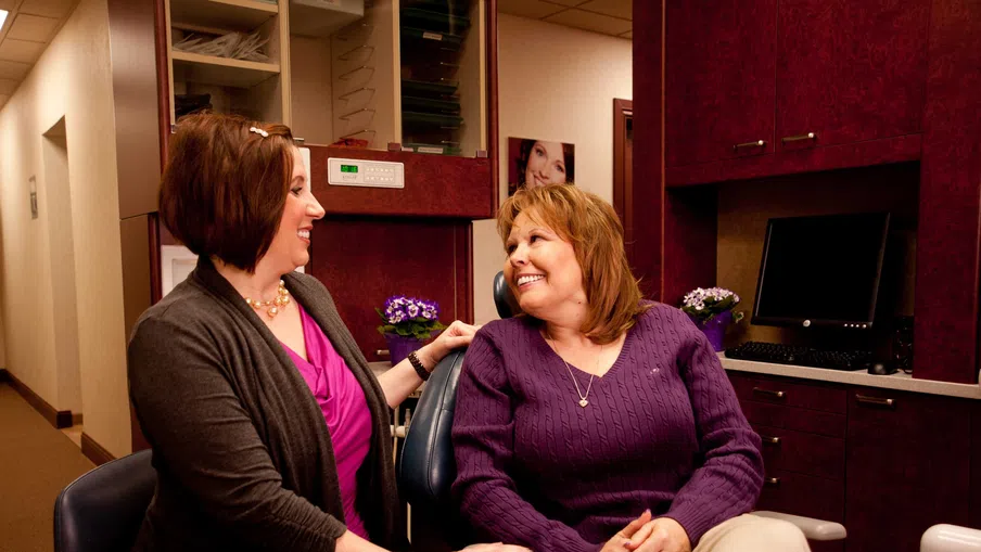 Smiling woman sitting in dental chair talking with female dental professional in clinic setting