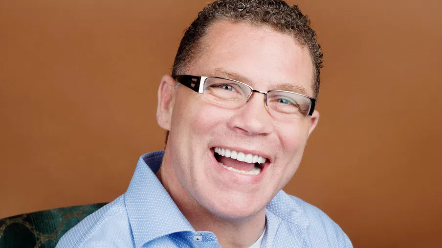 Smiling man with curly hair and glasses wearing a light blue shirt against a brown background