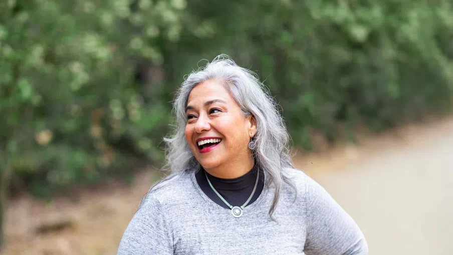 Smiling middle-aged woman with gray hair in a gray sweater outdoors on a path with greenery background