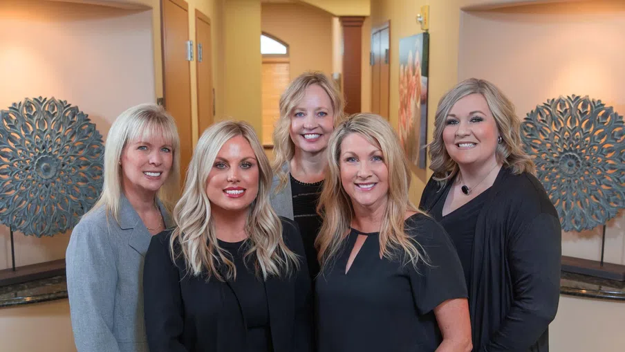 Five professional women posing and smiling in an office hallway with decorative wall art behind them.