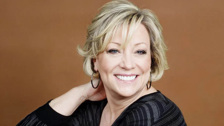Smiling middle-aged woman with short blonde hair wearing hoop earrings and a black striped top on brown background