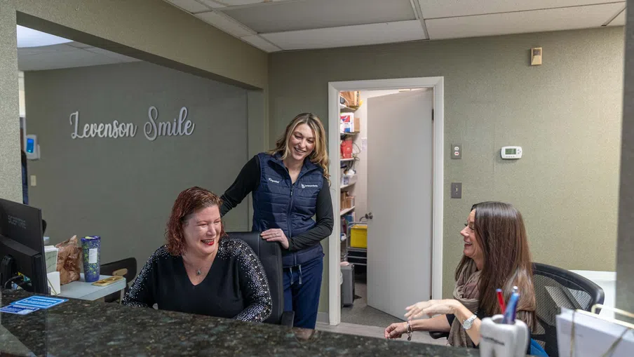 Three women smiling and chatting at the reception desk inside Levenson Smile dental office.