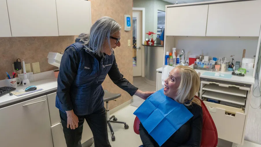 Dentist smiling and talking with female patient seated in dental chair wearing a blue bib in clinic.