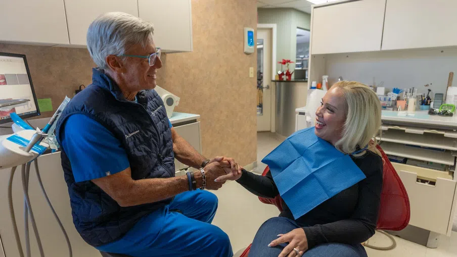 Dentist in blue scrubs holding patient hands and smiling in a modern dental office during consultation.