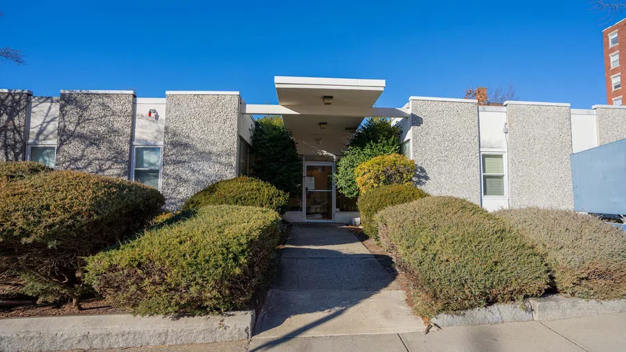 Modern one-story building entrance with concrete pathway and neatly trimmed bushes under clear blue sky.