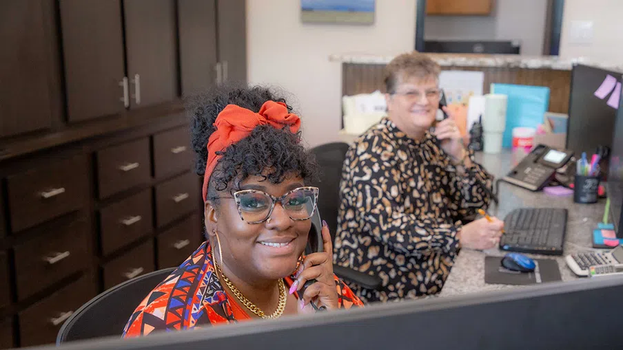 Two women smiling on phones while working at desks with computers and office supplies in a friendly workspace.