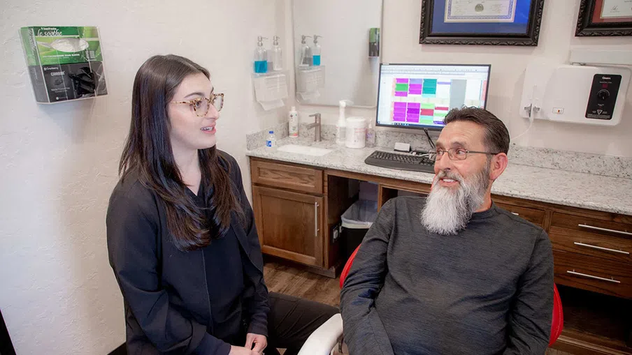 Dentist consulting with male patient in modern dental office with computer and certificates on wall.