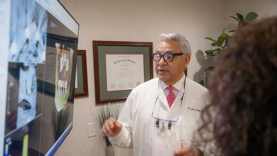 Dentist explaining dental X-rays on screen to a patient in a consultation room with framed diplomas.