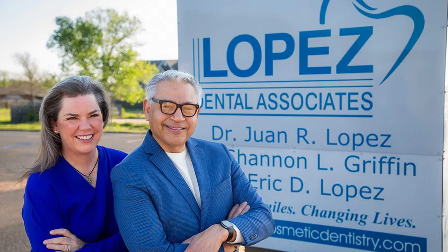 Smiling dental professionals standing in front of Lopez Dental Associates outdoor sign on sunny day