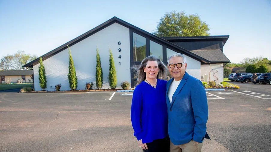 Smiling couple standing outside modern white building with address 6941 on a sunny day in parking lot.