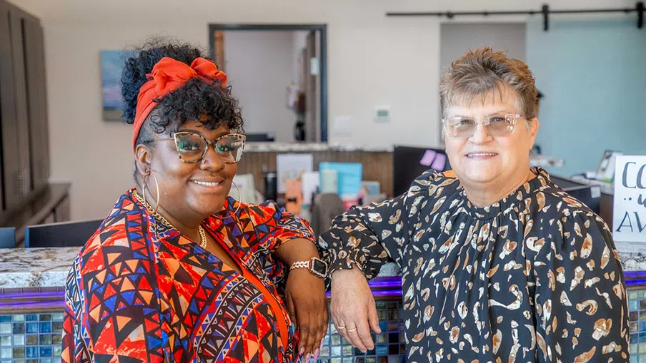 Two smiling women standing and leaning on a mosaic tiled counter in a brightly lit interior space.