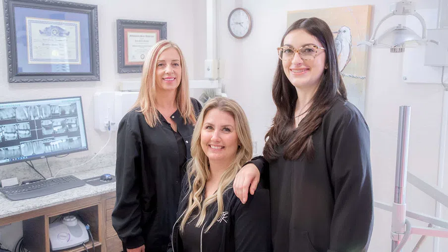 Three female dental professionals in black uniforms smiling inside a bright dental office with certificates and equipment.