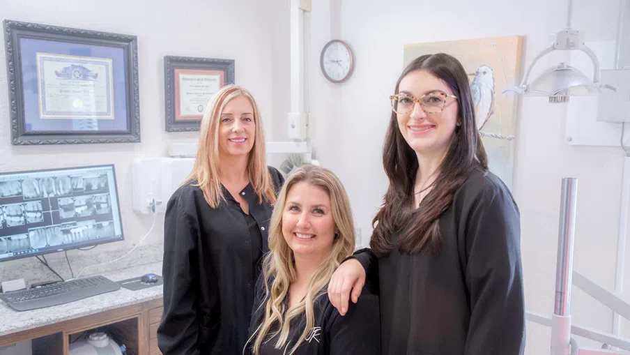 Three female dental professionals in black uniforms smiling inside a bright dental office with certificates and equipment.